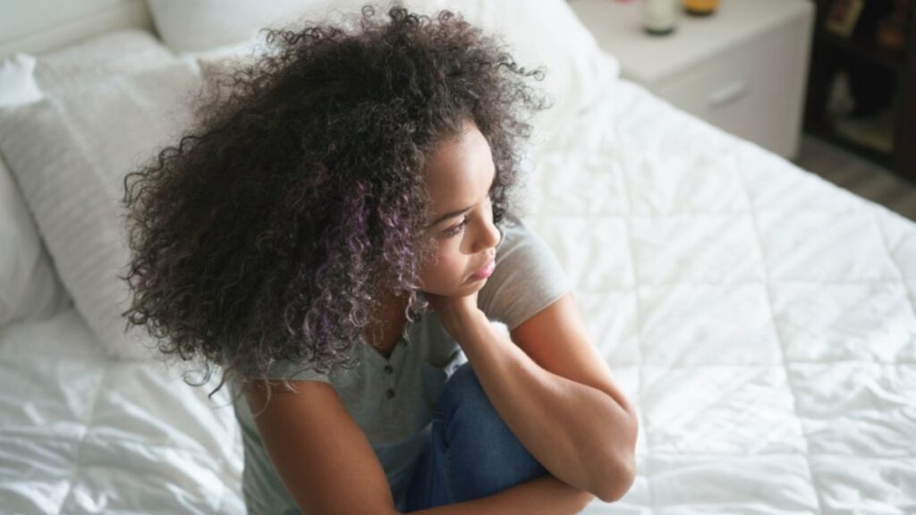 A young woman seated on a bed, with her hands on her chin, deep in thought about building a meaningful life despite feeling lost.
