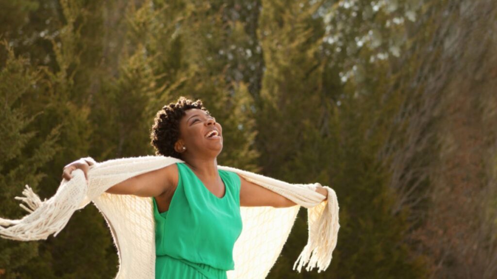 A woman in a flowing green dress smiles with outstretched arms on a hillside at sunset, representing joy and fulfillment.